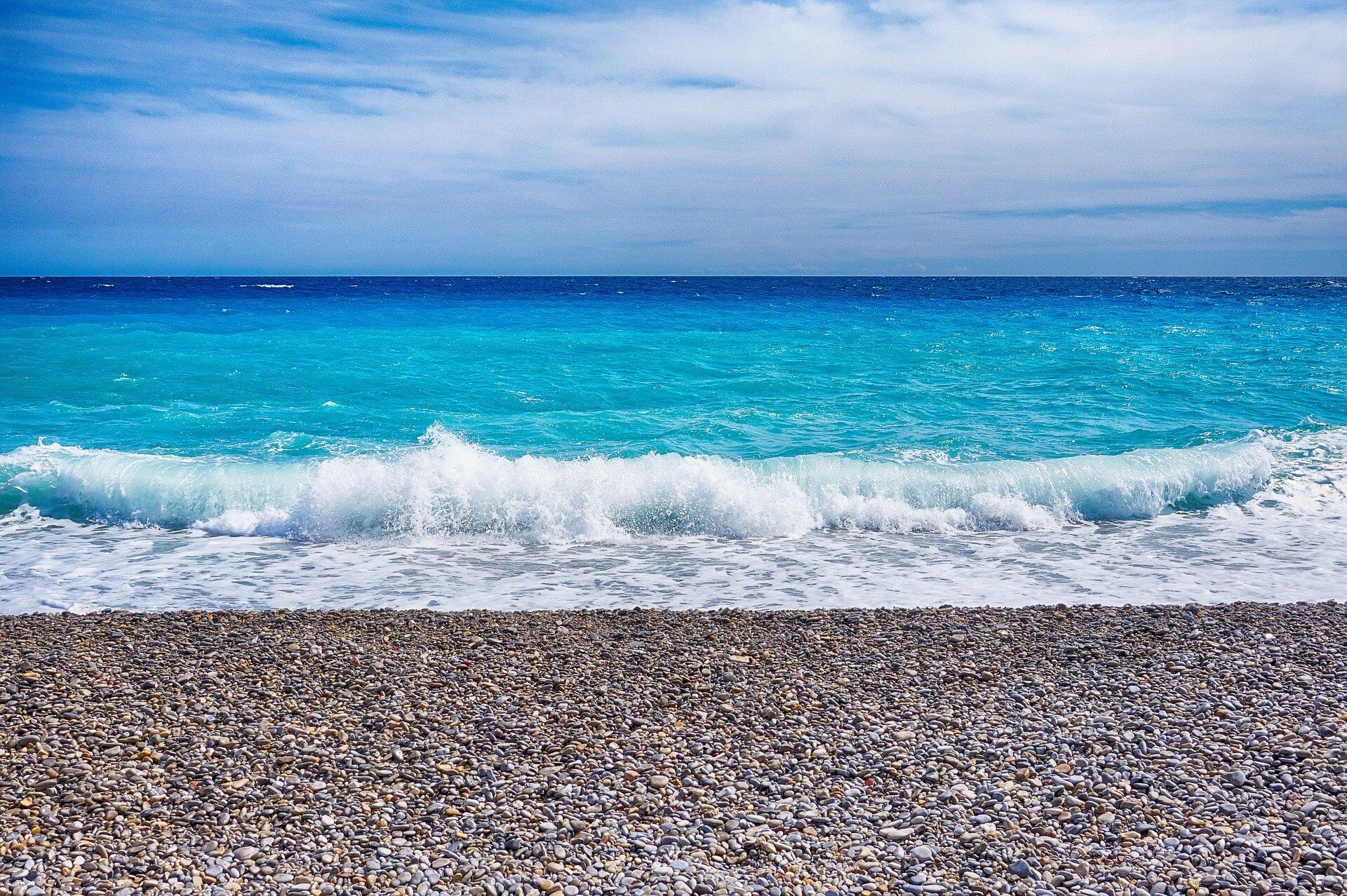Bright blue Mediterranean waves reaching a pebble shore beneath an open sky.